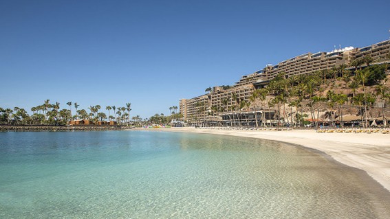 Ruhige Badebucht mit klarem Wasser, heller Sandstrand und terrassenförmig gebaute Hotelanlagen am Hang.